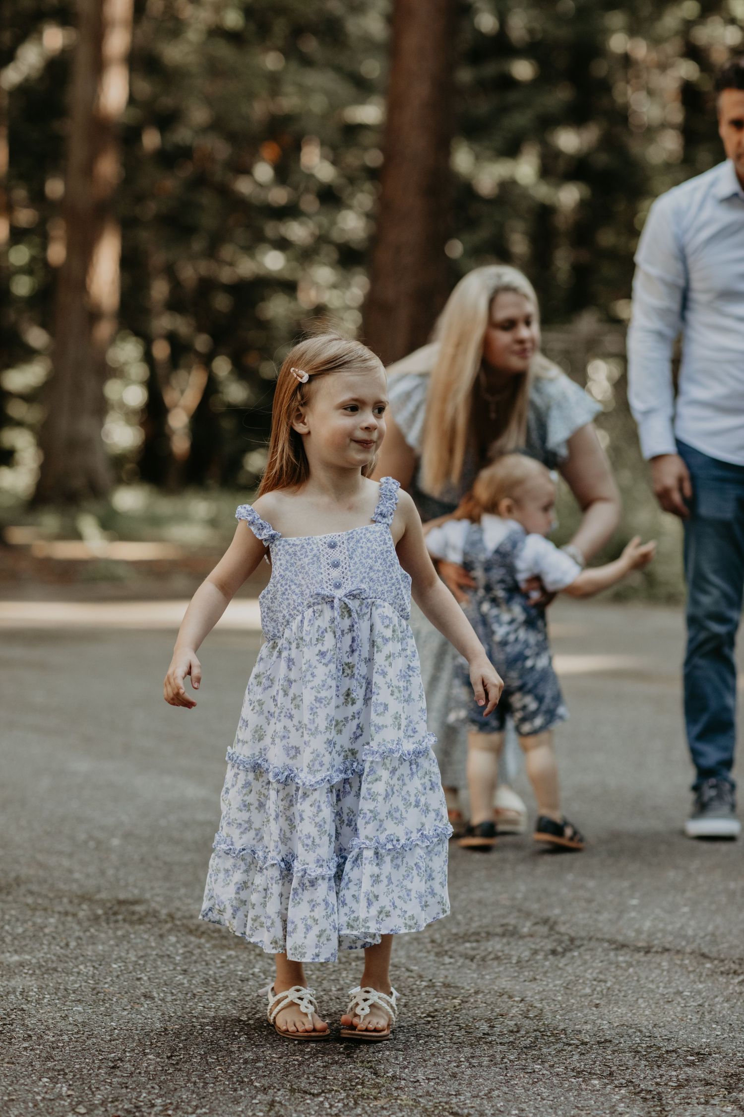 Young girl in blue floral dress walks happily on a forest path as family gathers in the background.