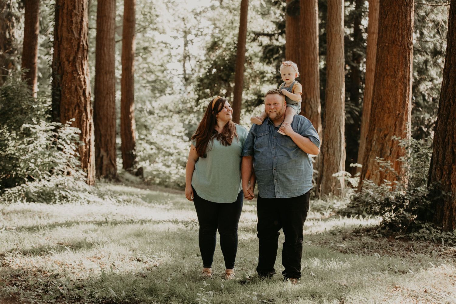 Three people walk together on a sunlit forest path among tall redwood trees on a summer day.