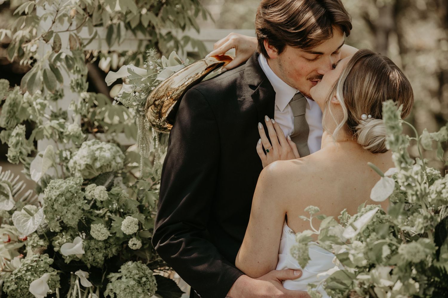 A romantic wedding couple embraces among lush greenery and white flowers in a dreamy outdoor garden setting.
