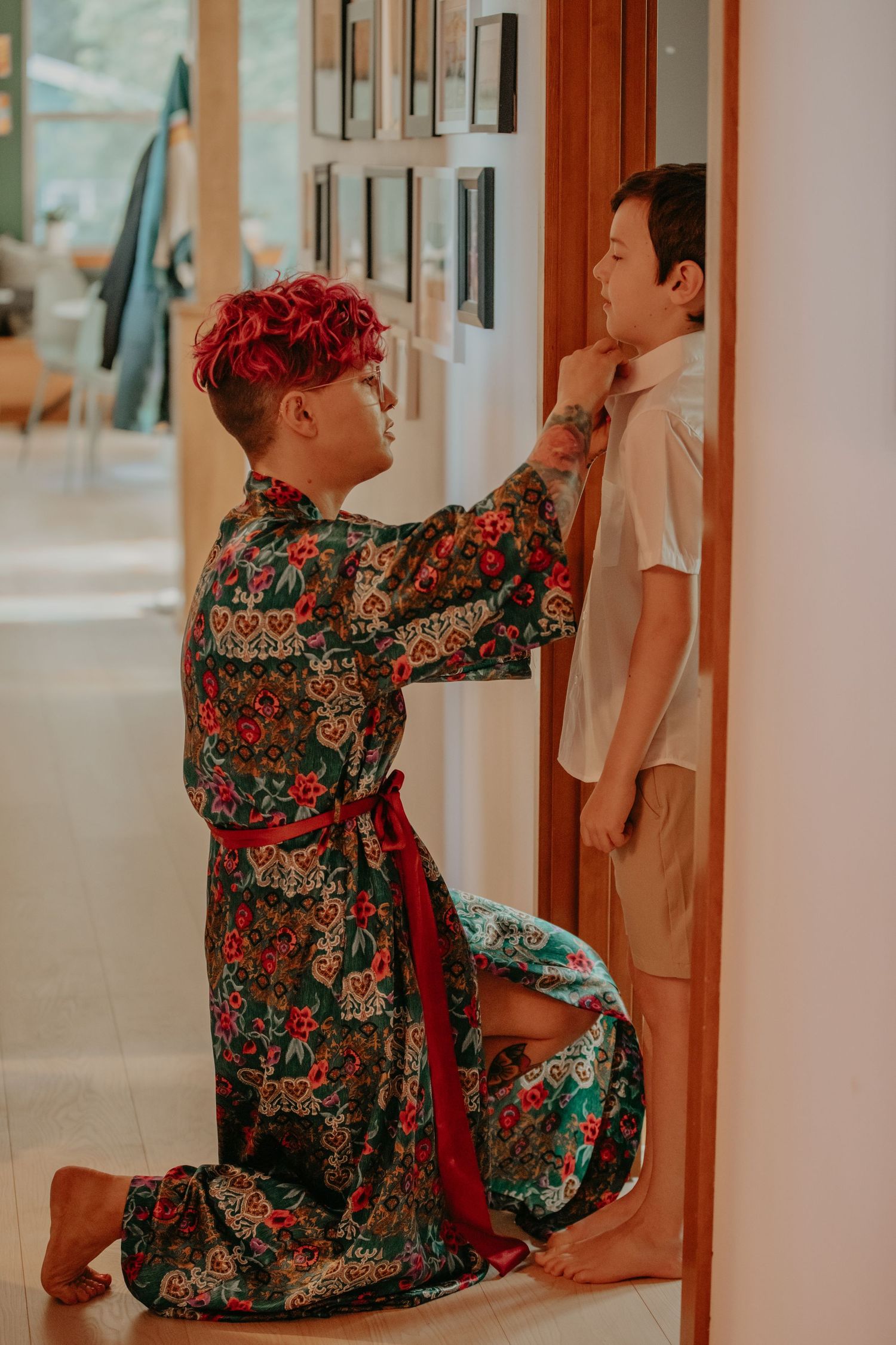 A person in a vibrant floral kimono with red hair adjusts someone's collar in a hallway.