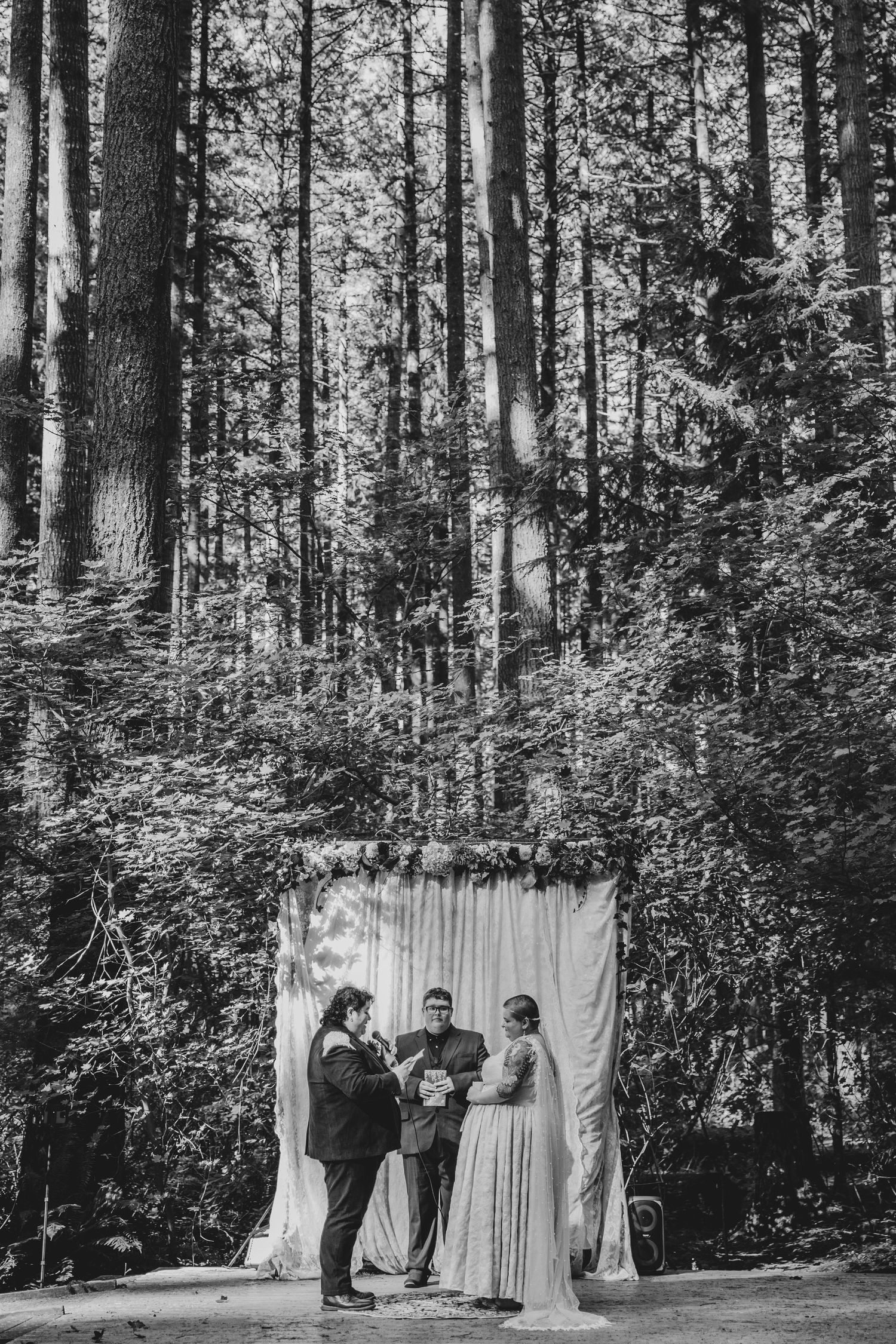 A romantic outdoor wedding ceremony takes place in a serene forest setting with white drapes hanging between tall trees.