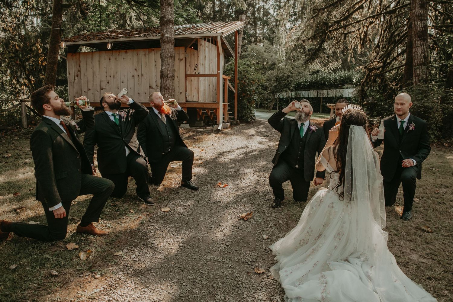 Wedding party having fun posing together on a rustic outdoor path with bride in flowing veil and groomsmen jumping.