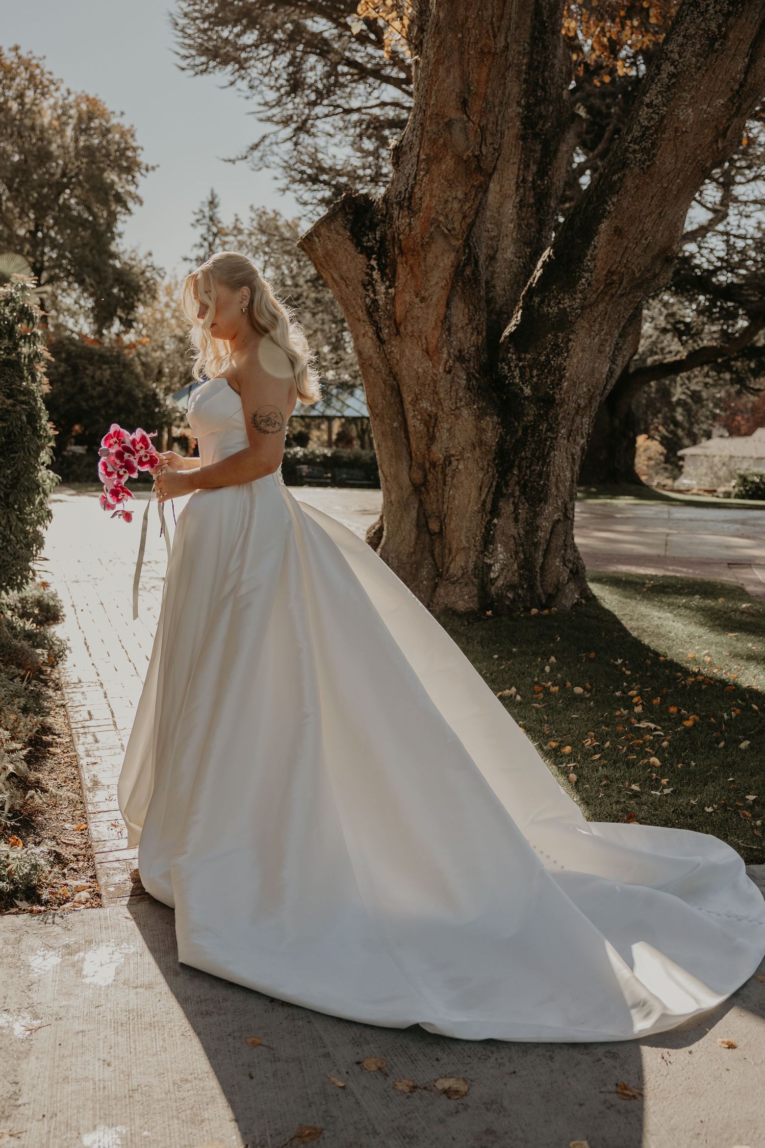 A bride in an elegant white ballgown wedding dress with a long train stands beside a large tree holding a pink bouquet.