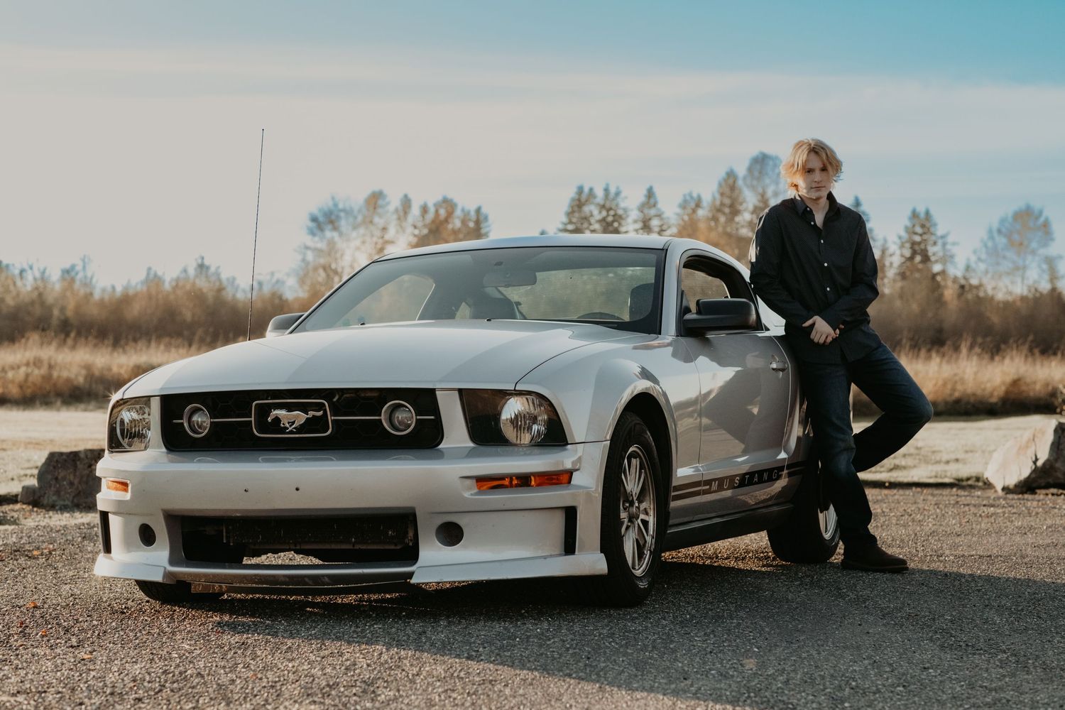 A silver Ford Mustang GT parked on a rural road with trees in the background on a sunny day.