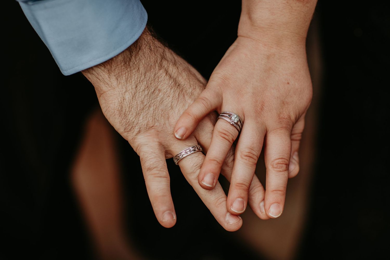 Close-up view of two hands wearing wedding rings touching gently against a dark background.