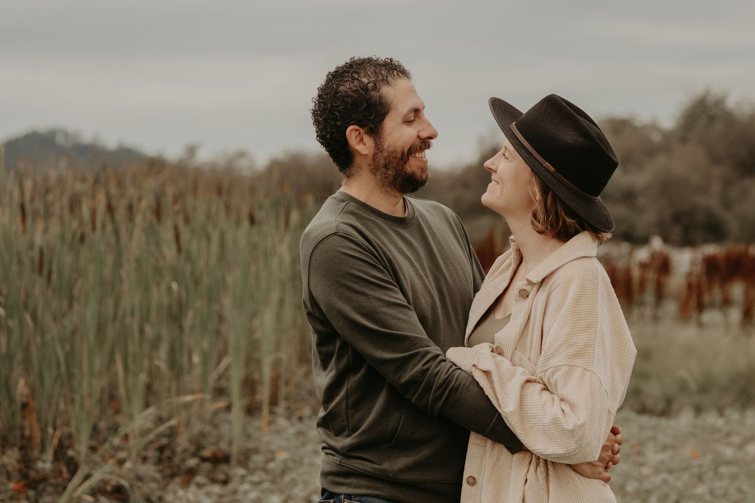 Couple embraces lovingly in rustic field with grazing horses in background during golden hour.