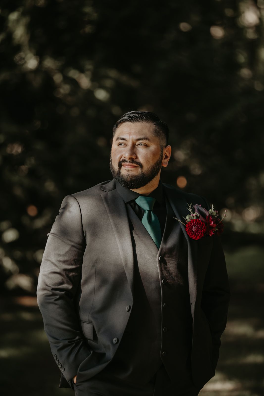 A groom in a charcoal suit and emerald tie poses for portraits in an outdoor setting with bokeh lighting effects.