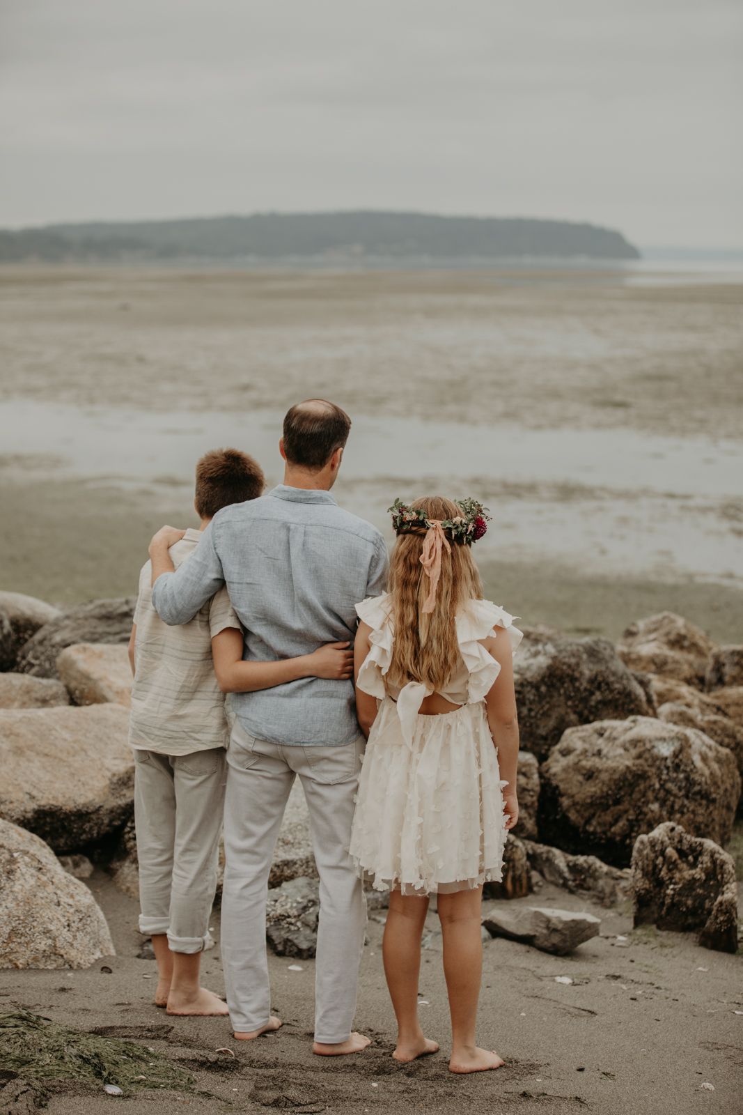 Family in coordinated neutral outfits standing together on a rocky beach looking at the ocean view.