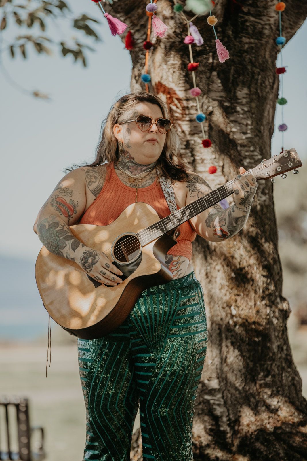 Musician in green sequined pants plays acoustic guitar under flowering tree branches.