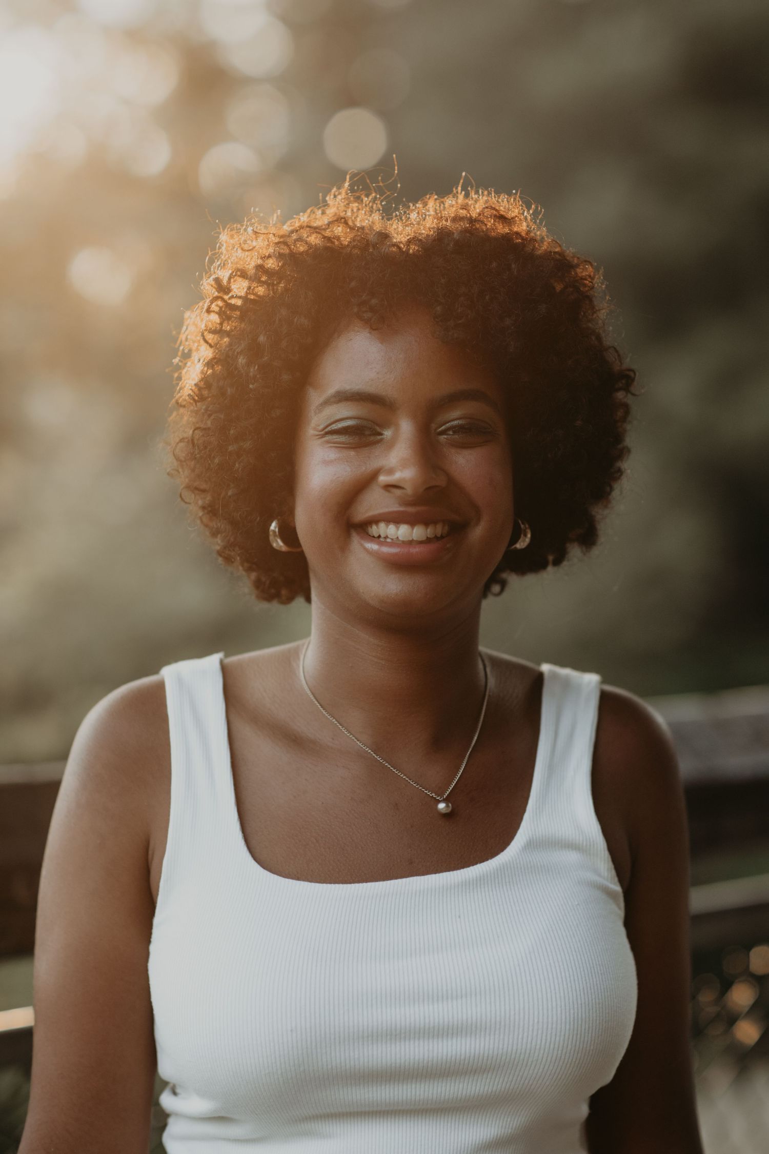 Smiling person in a white tank top with natural curly hair and necklace in warm sunset lighting.