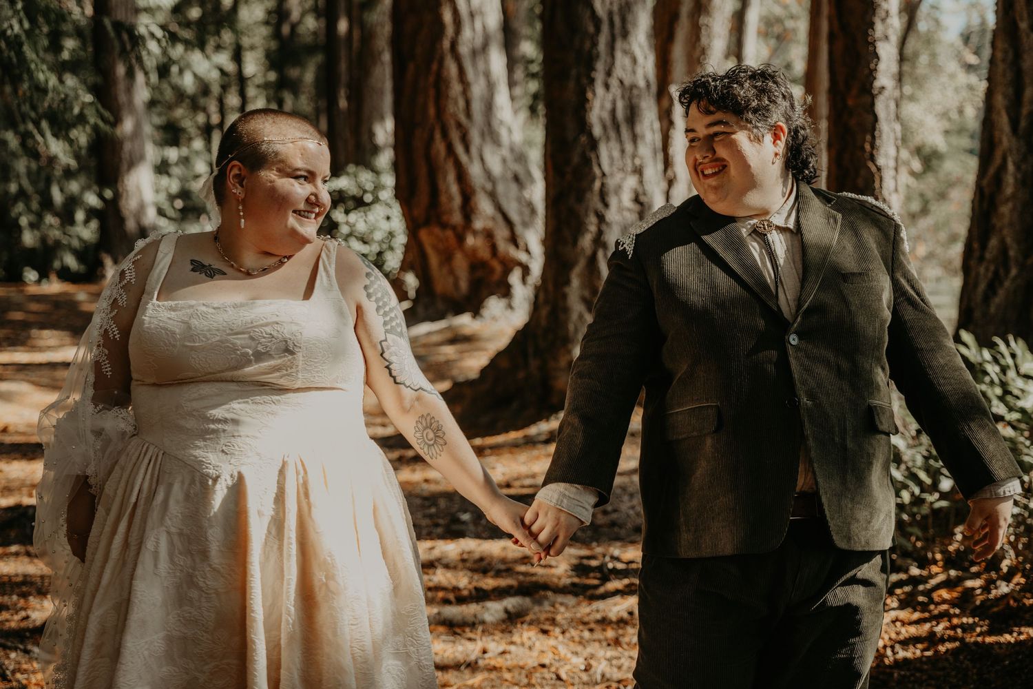 A couple holds hands walking through a sunlit forest, one in a white wedding dress and one in a dark suit.