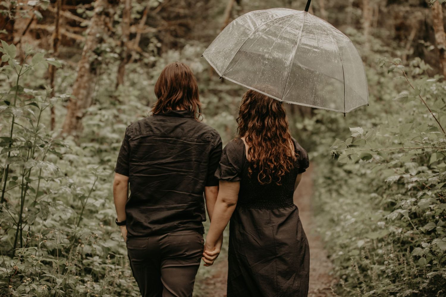 Two figures sharing an umbrella walk together through lush greenery on a moody day.