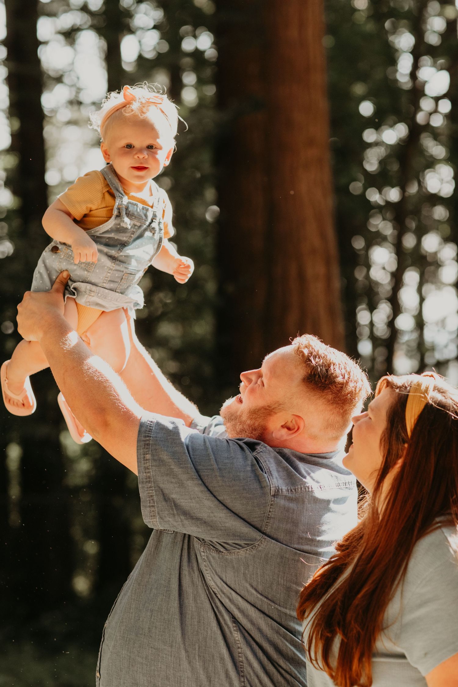 Parents playing with smiling baby in sunlit forest setting with tall trees in background.