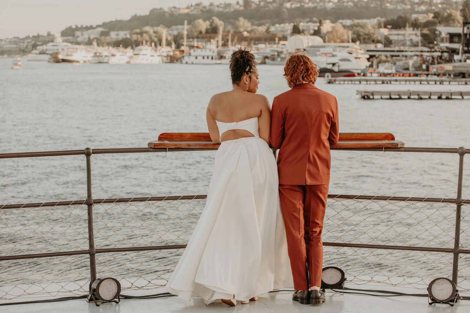 A couple in wedding attire stands at a waterfront railing overlooking a harbor on their special day.