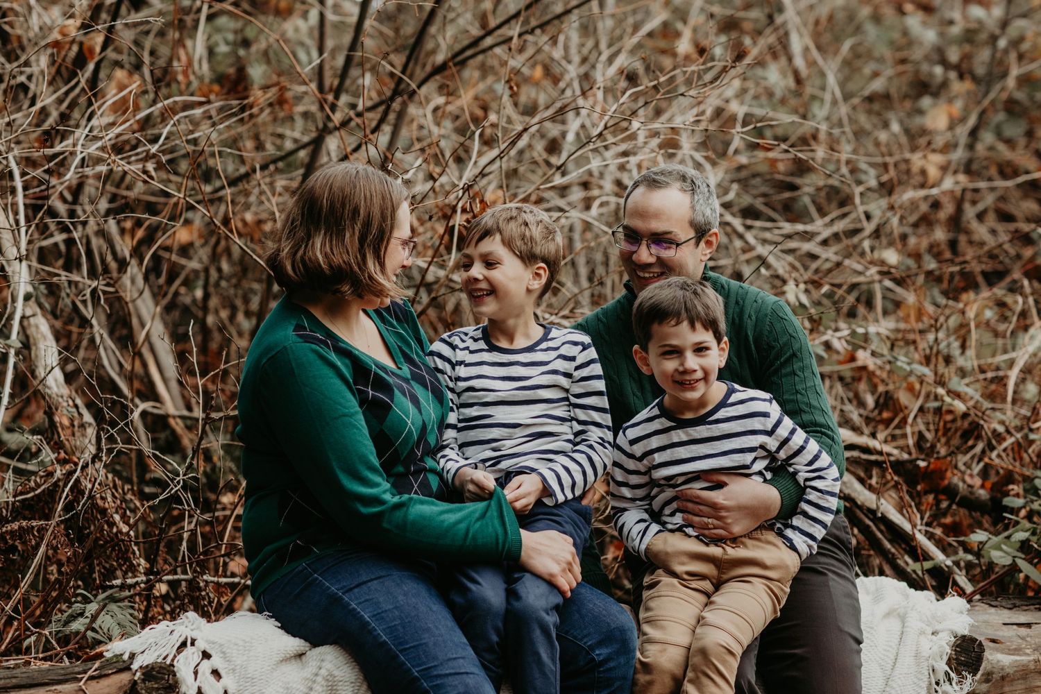 Family wearing matching green sweaters sits together on outdoor bench surrounded by autumn foliage in natural setting.