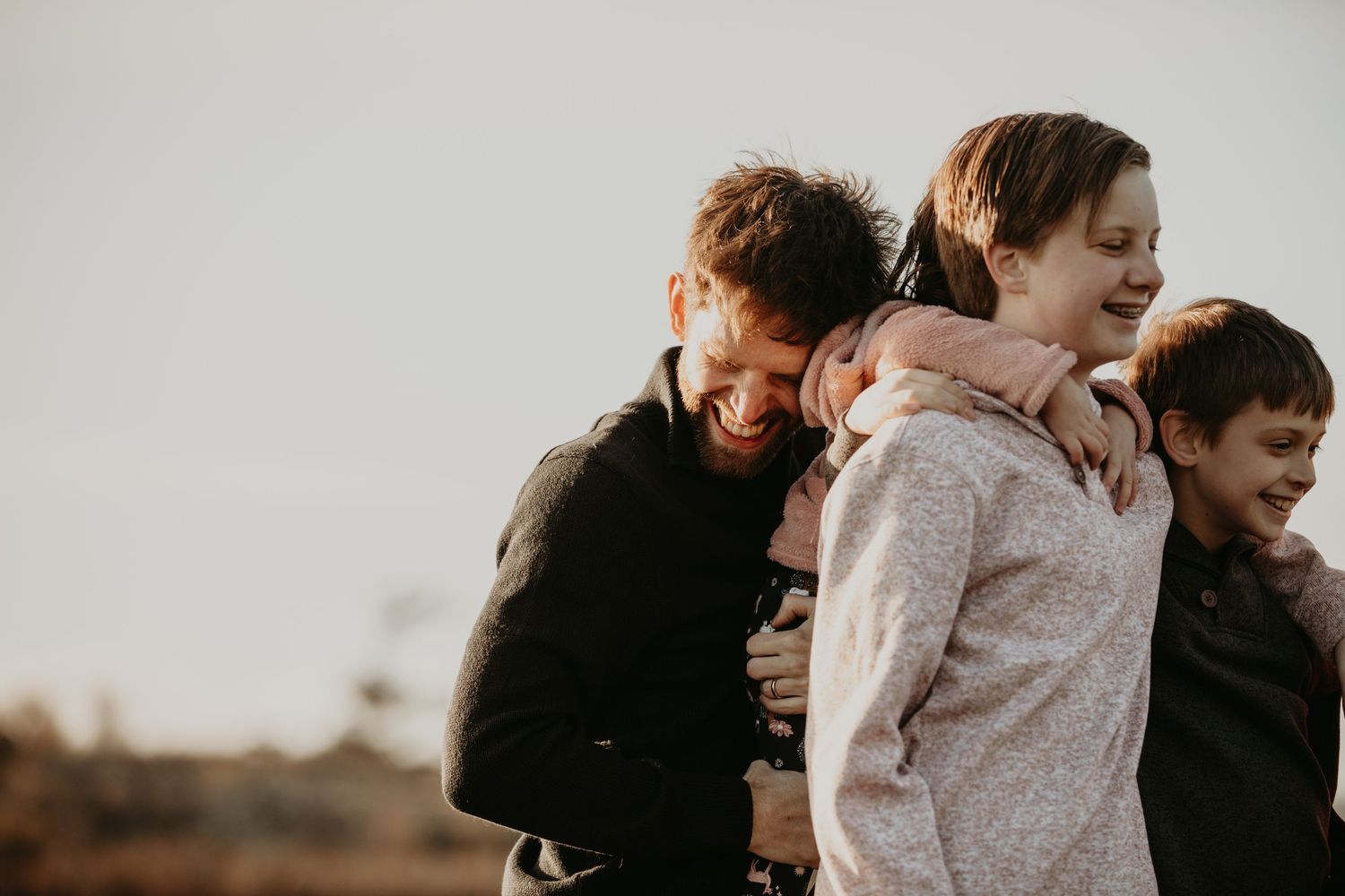 Three people hugging and laughing together outdoors at sunset, wearing dark and light colored sweaters.