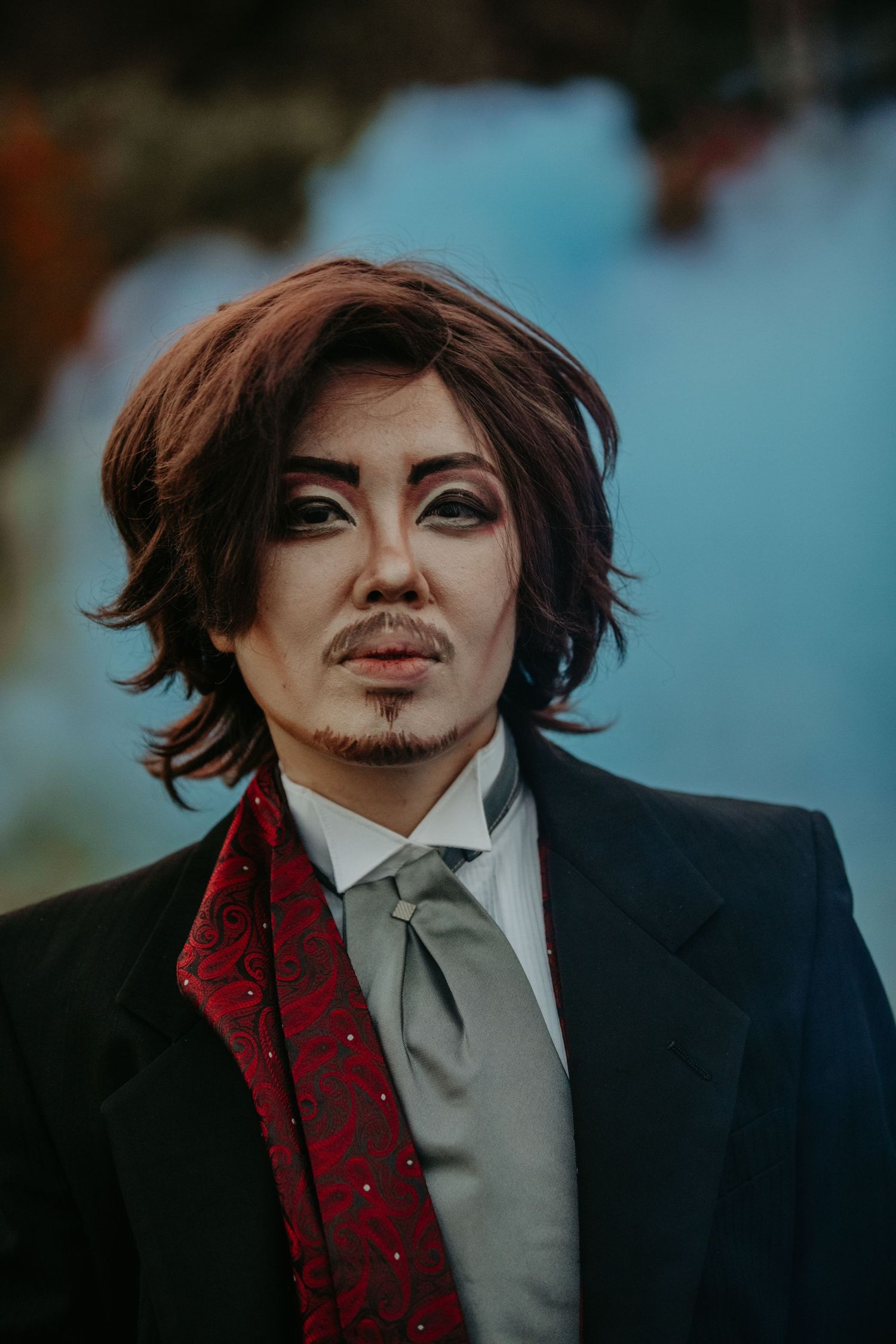Person with angular features wearing black suit and red polka dot tie poses for dramatic portrait in moody lighting.