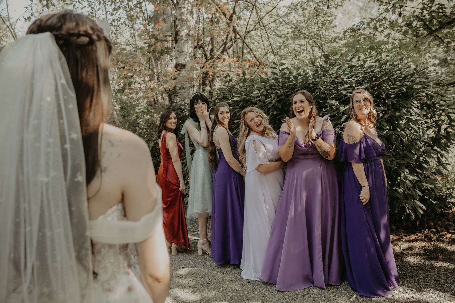 Wedding party members in purple and lavender dresses gather outdoors for group photos in a garden setting.