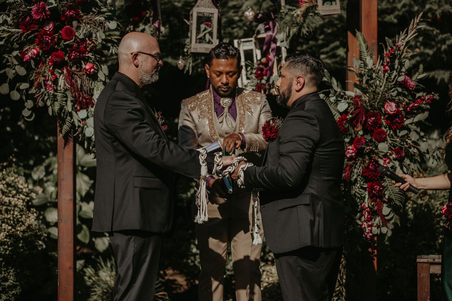 Two people in dark suits share a romantic moment under a floral arch at an outdoor wedding ceremony.