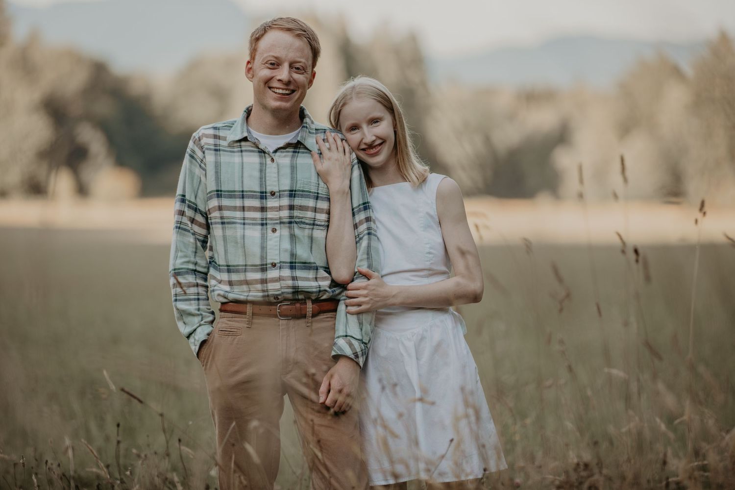 A couple stands close together in a golden wheat field during golden hour wearing rustic casual attire.