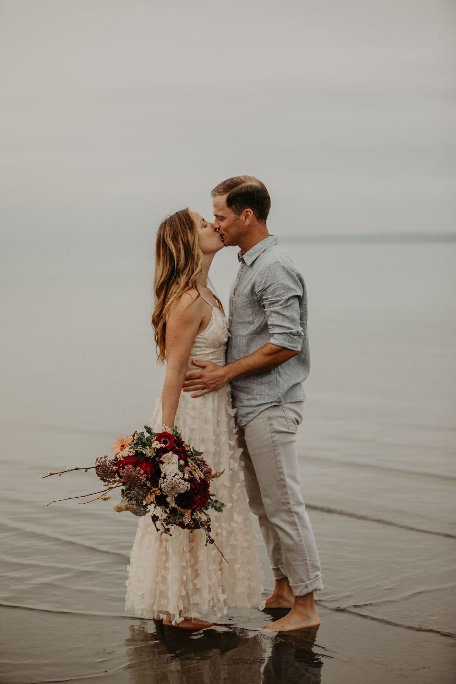 A couple shares a romantic moment on a beach at sunset with the bride holding a burgundy and blush floral bouquet.