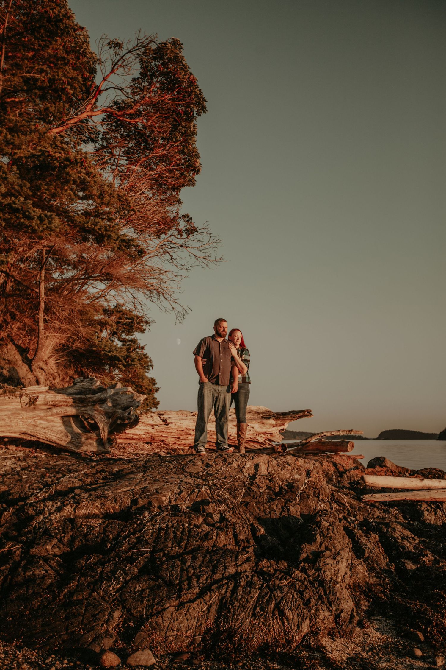 A couple stands on a rocky coastal cliff at sunset with autumn-colored trees and ocean views in the background.