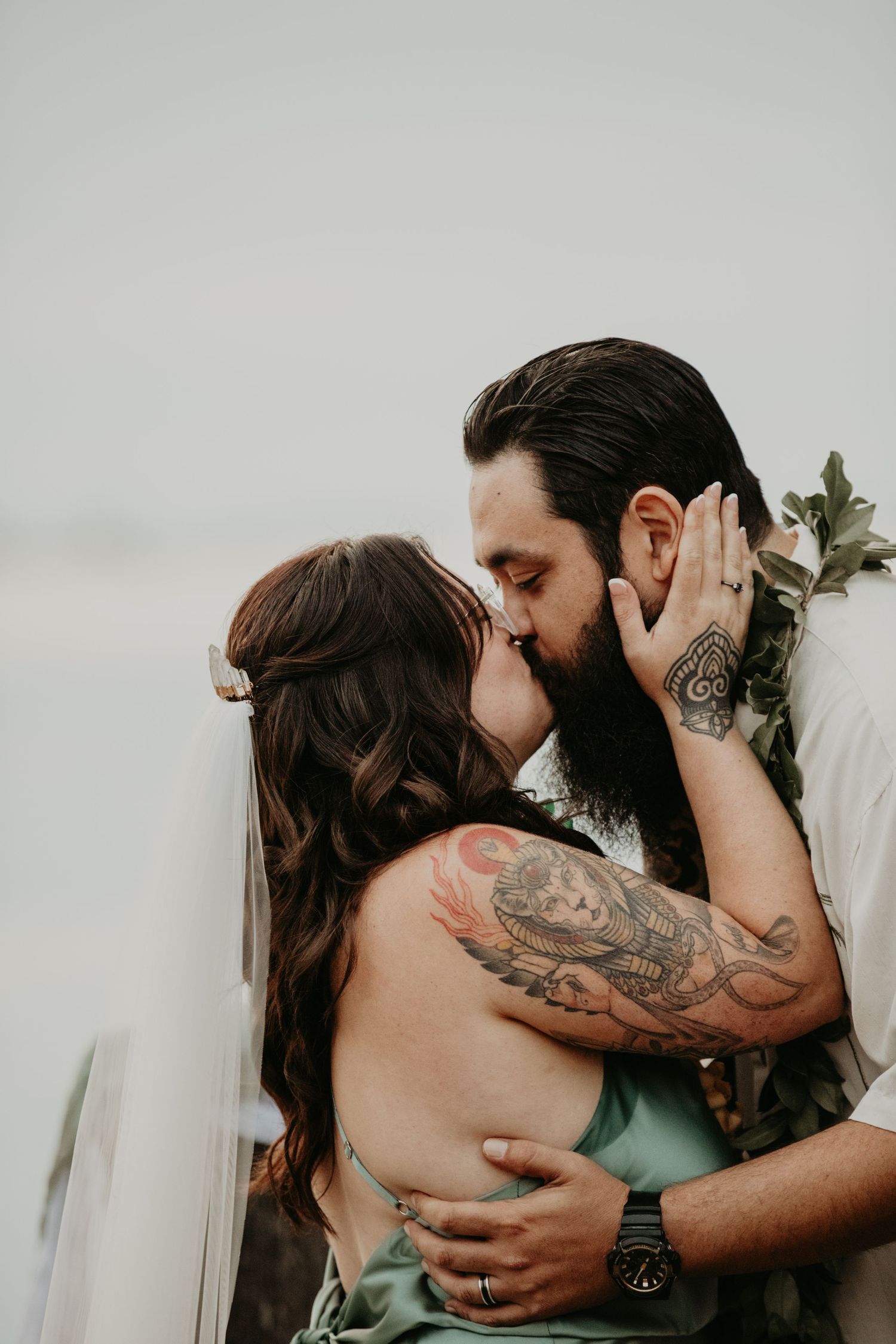 A romantic couple sharing a passionate kiss while embracing, with visible tattoos and wedding attire against a light background.