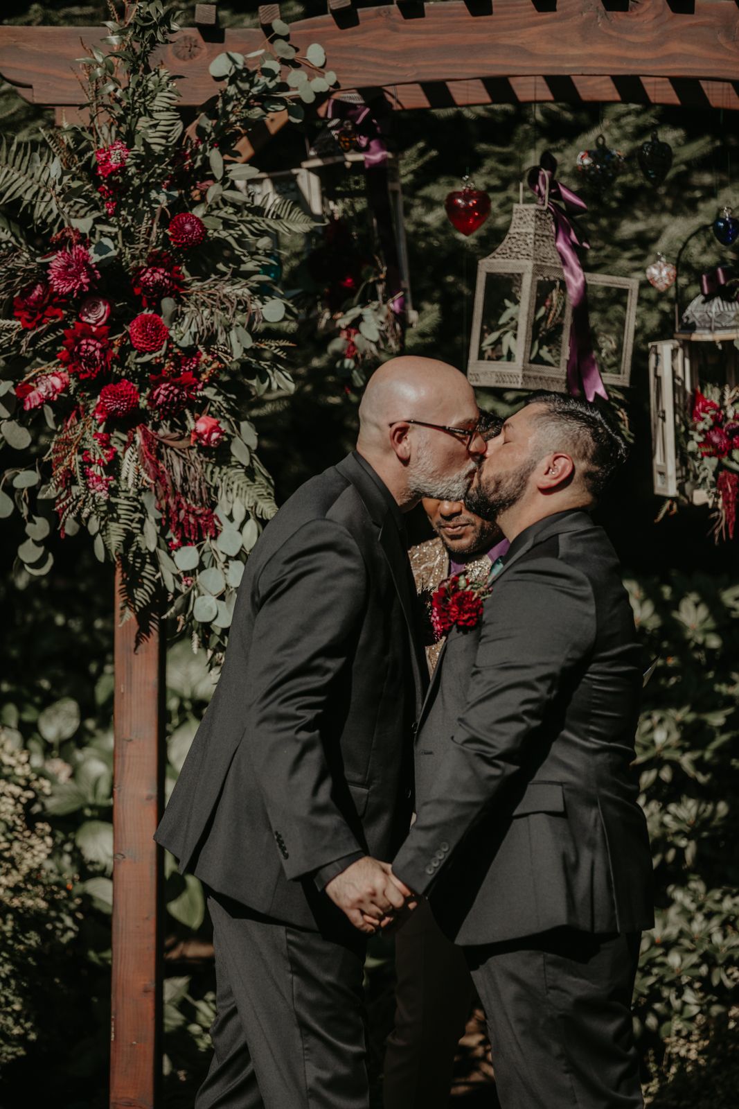A couple shares an intimate moment under a wooden pergola decorated with vibrant red flowers during a wedding ceremony.
