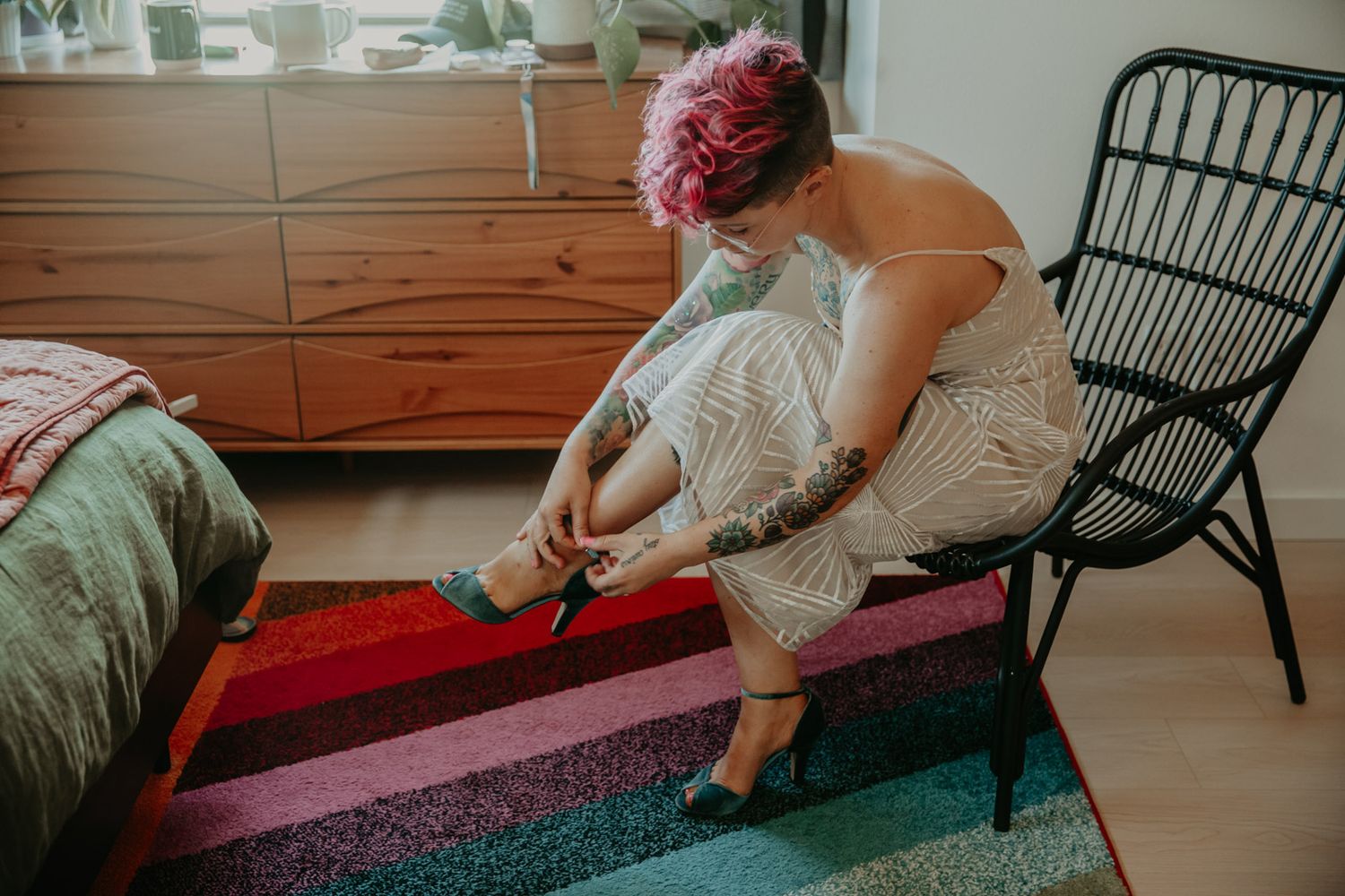 Person with pink hair wearing a white lace dress fastens strappy heels while sitting on a black rattan chair.