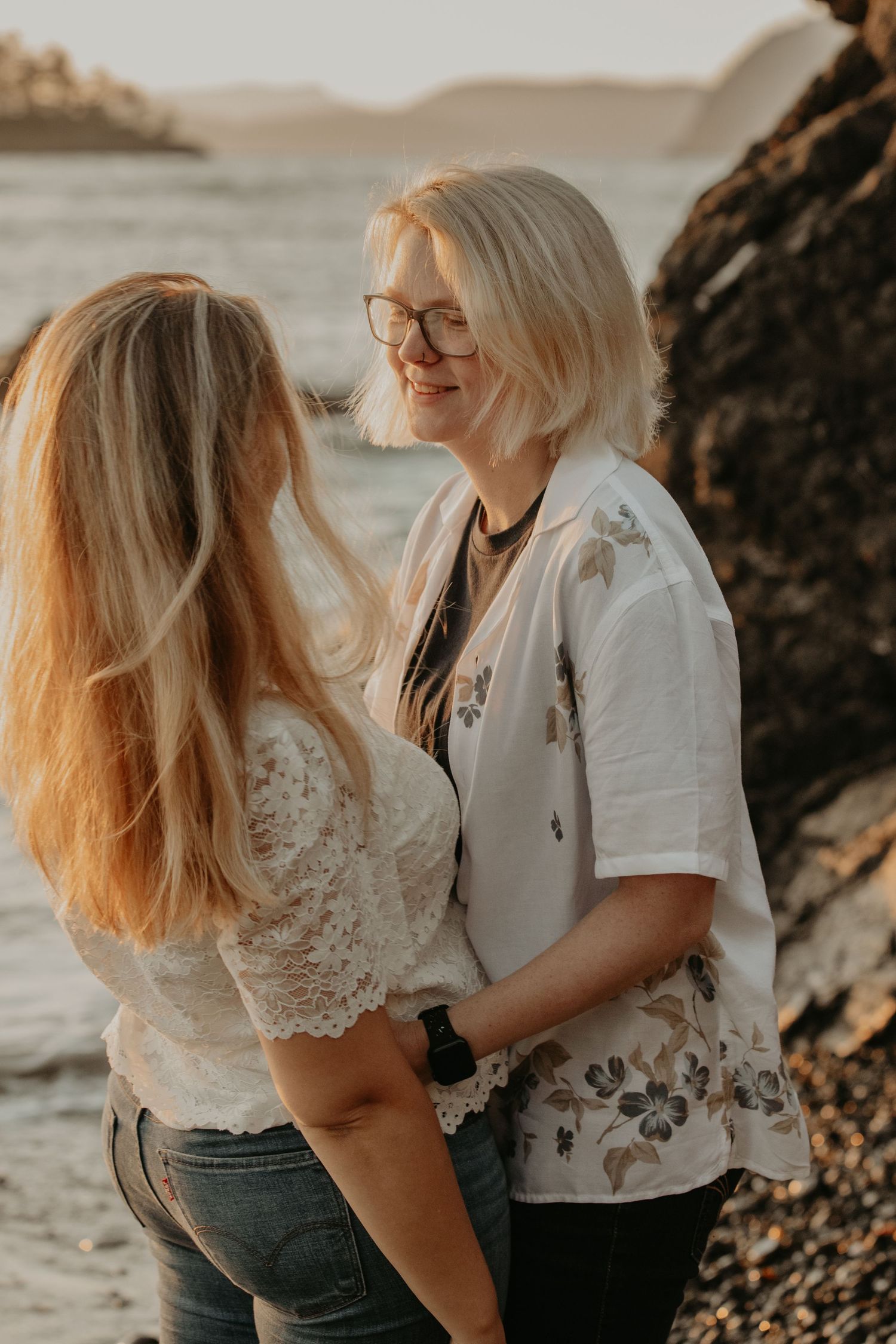 Couple sharing tender moments on a rocky beach at sunset in matching white casual outfits.