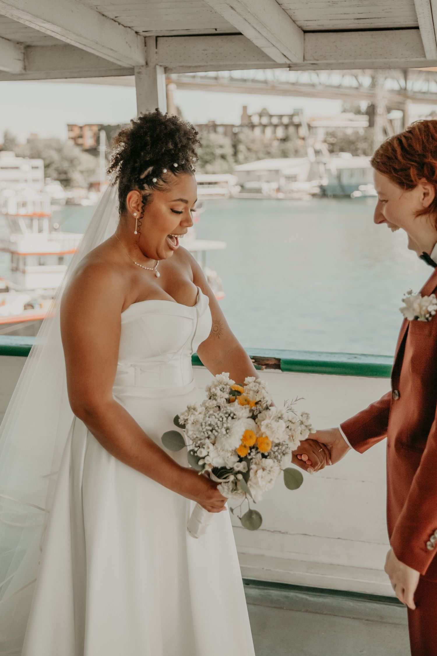 Wedding party members share candid moments on a waterfront deck with boats in the background.