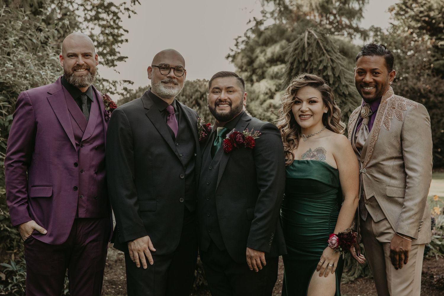 Wedding party members in formal attire pose together outdoors against a natural backdrop.