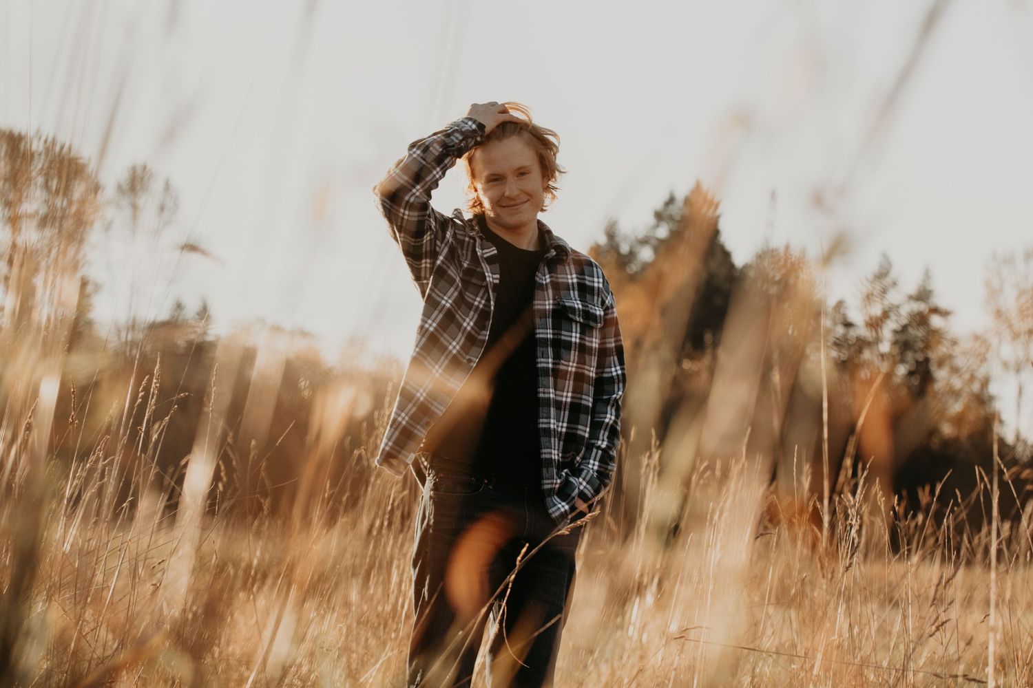 Person in plaid shirt poses in golden field of tall grass during sunset with warm autumn lighting.