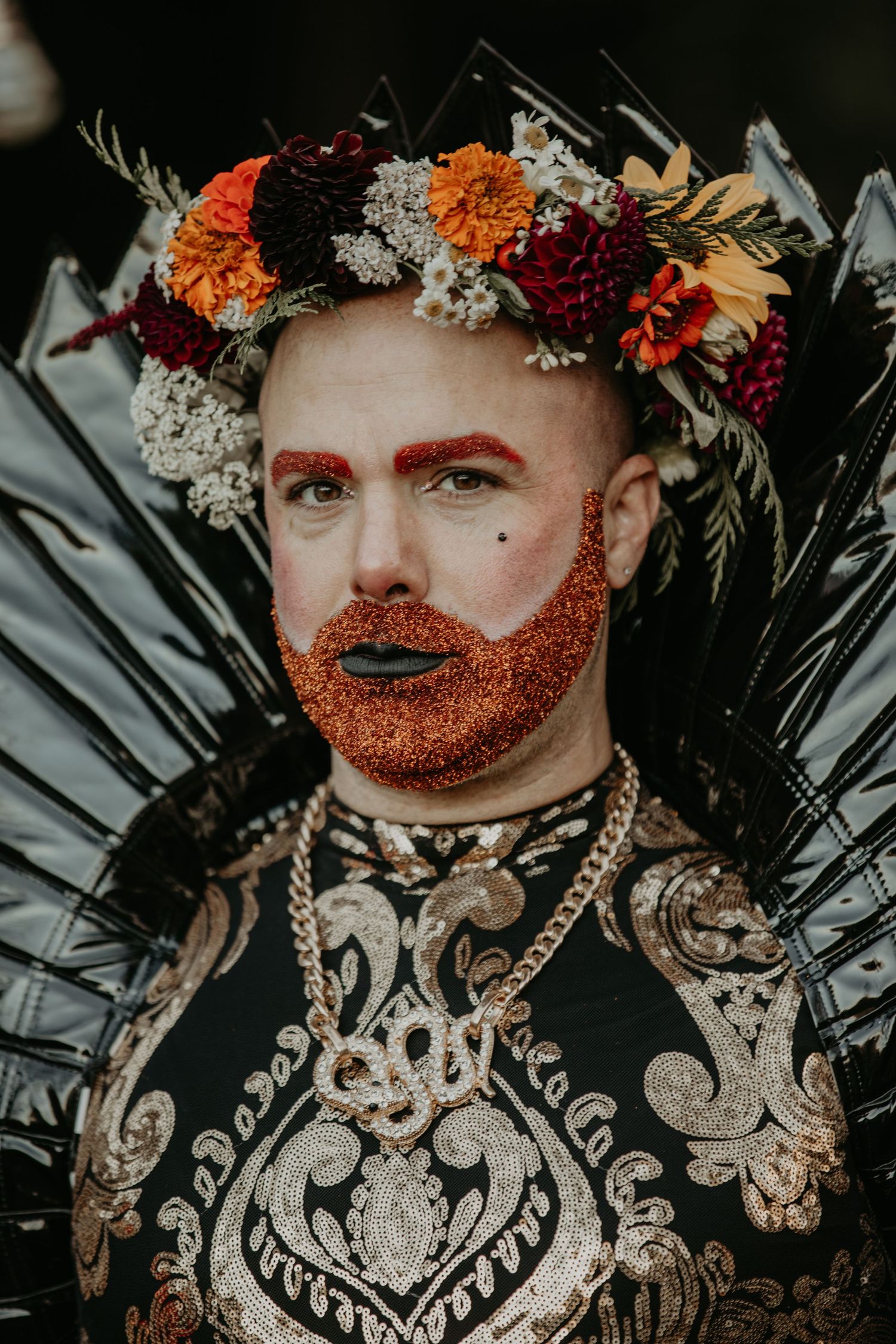 A portrait wearing an ornate black patterned garment with orange glitter beard and floral crown of autumn colors.
