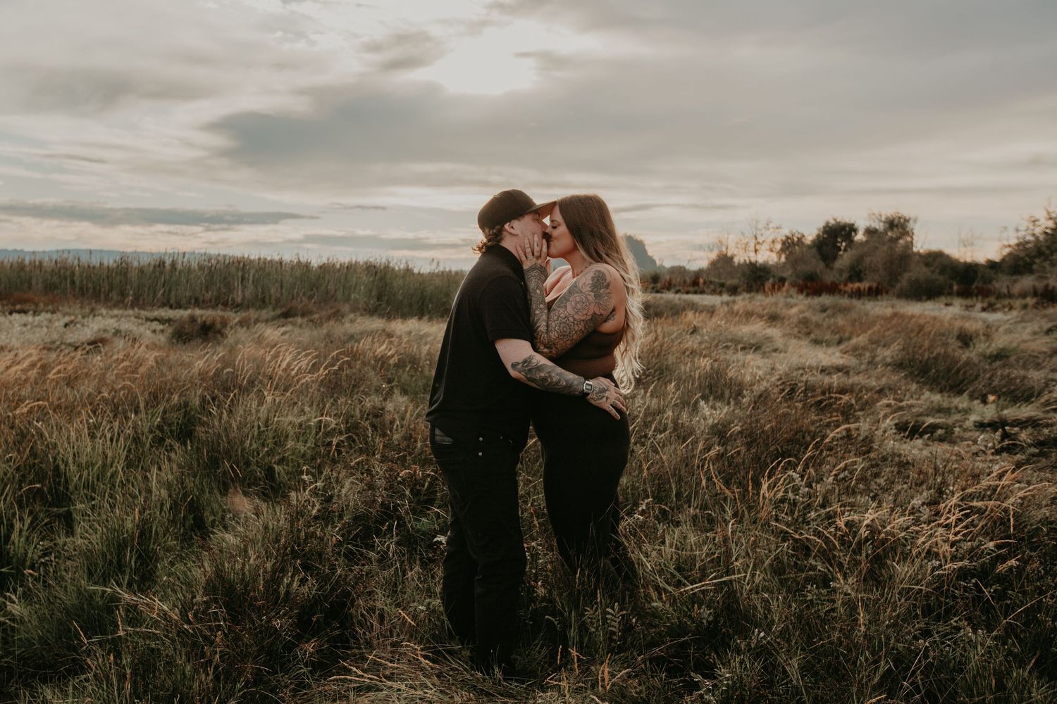 A couple shares an intimate embrace in a golden field at sunset with dramatic cloudy skies in the background.