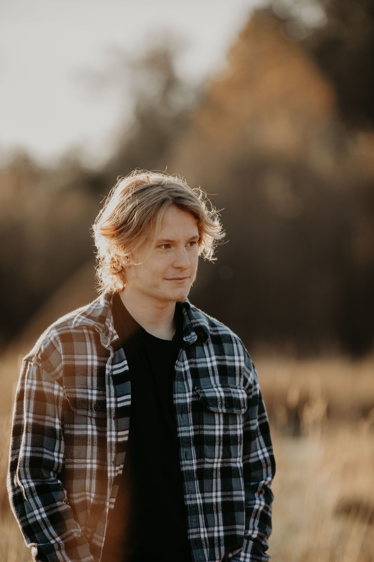 Person in plaid flannel shirt and black t-shirt standing in golden field at sunset with bokeh background.