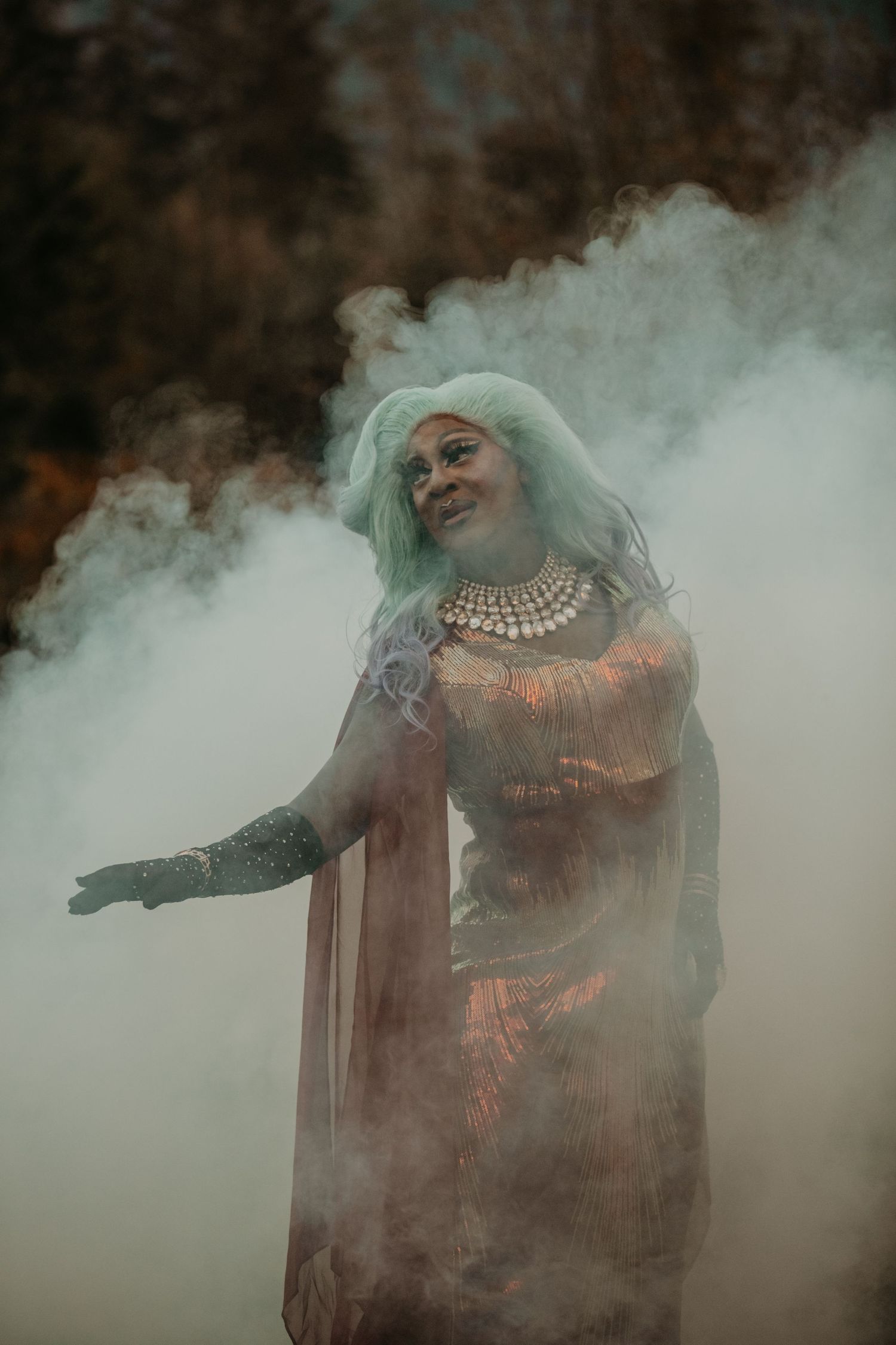 A figure in a glamorous gown and pearl necklace poses dramatically amidst swirling smoke effects in atmospheric lighting.
