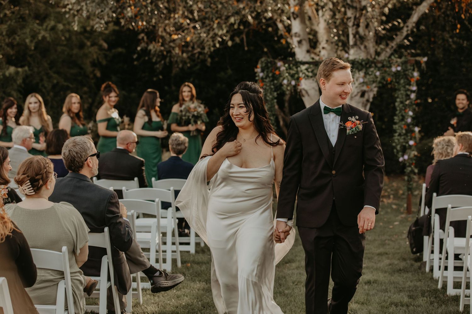 Wedding guests gather outdoors at ceremony seating in white chairs during sunset in wooded setting.