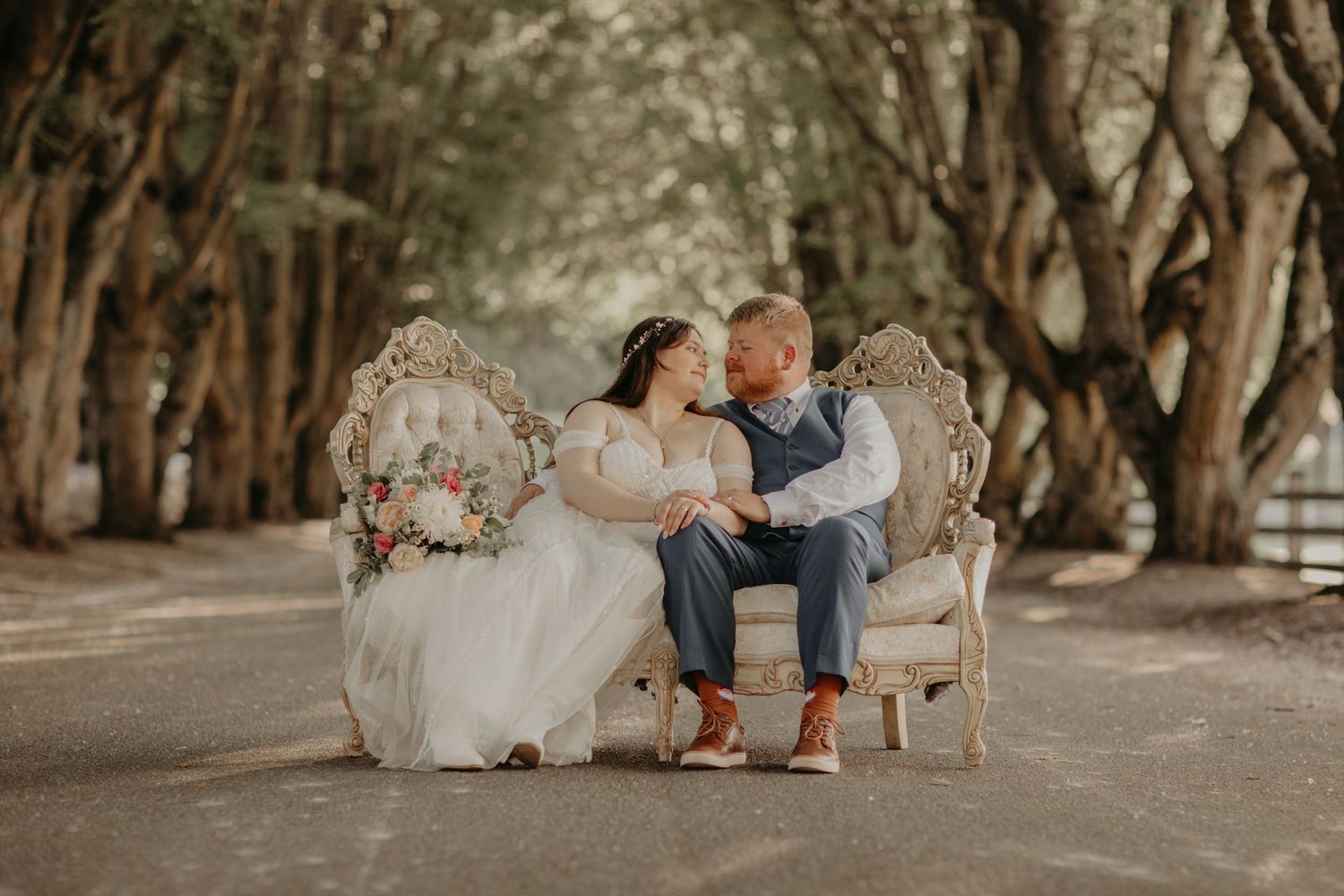 Bride and groom sitting on ornate vintage sofa on tree-lined path, sharing a romantic moment in wedding attire.