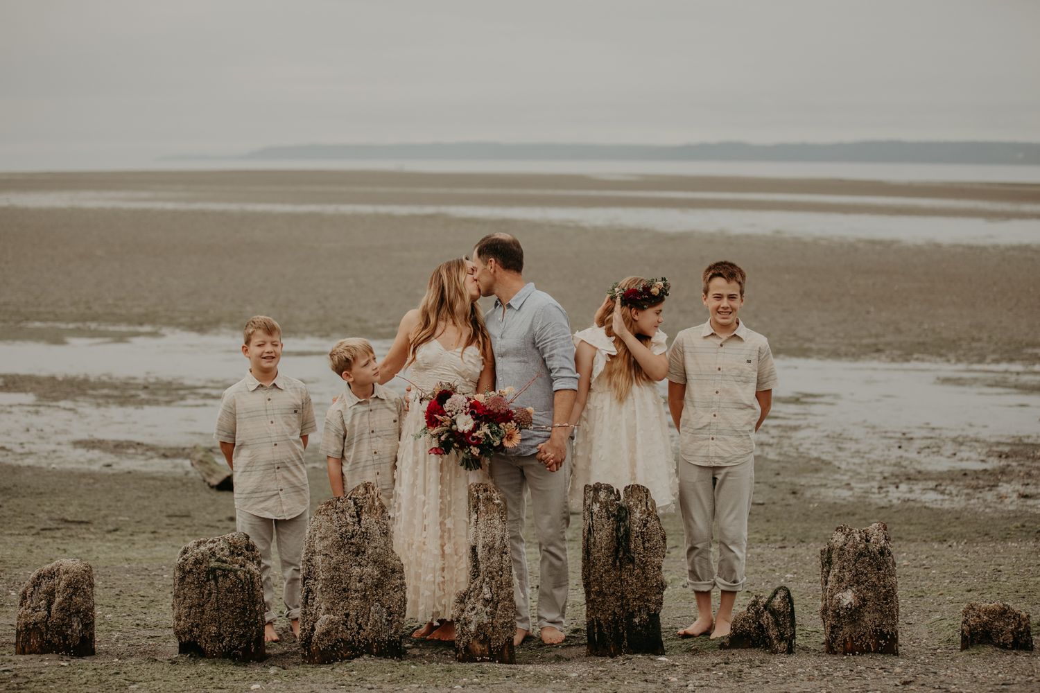A group of people in wedding attire pose together on a misty beach with waves crashing in the background.