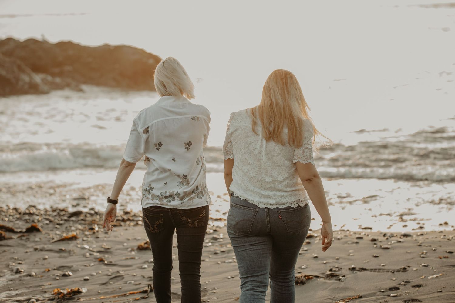 Two people walk hand in hand along a peaceful beach during sunset with waves gently rolling onto the shore.