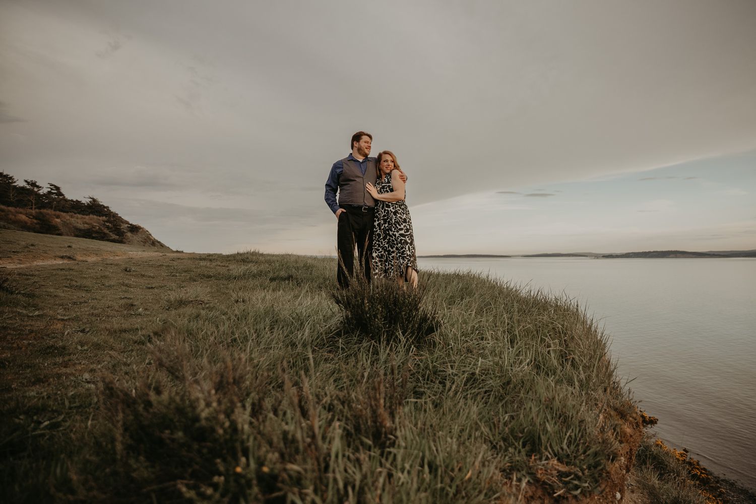 Couple embracing on a grassy coastal cliff at dusk with calm water and cloudy sky in the background.