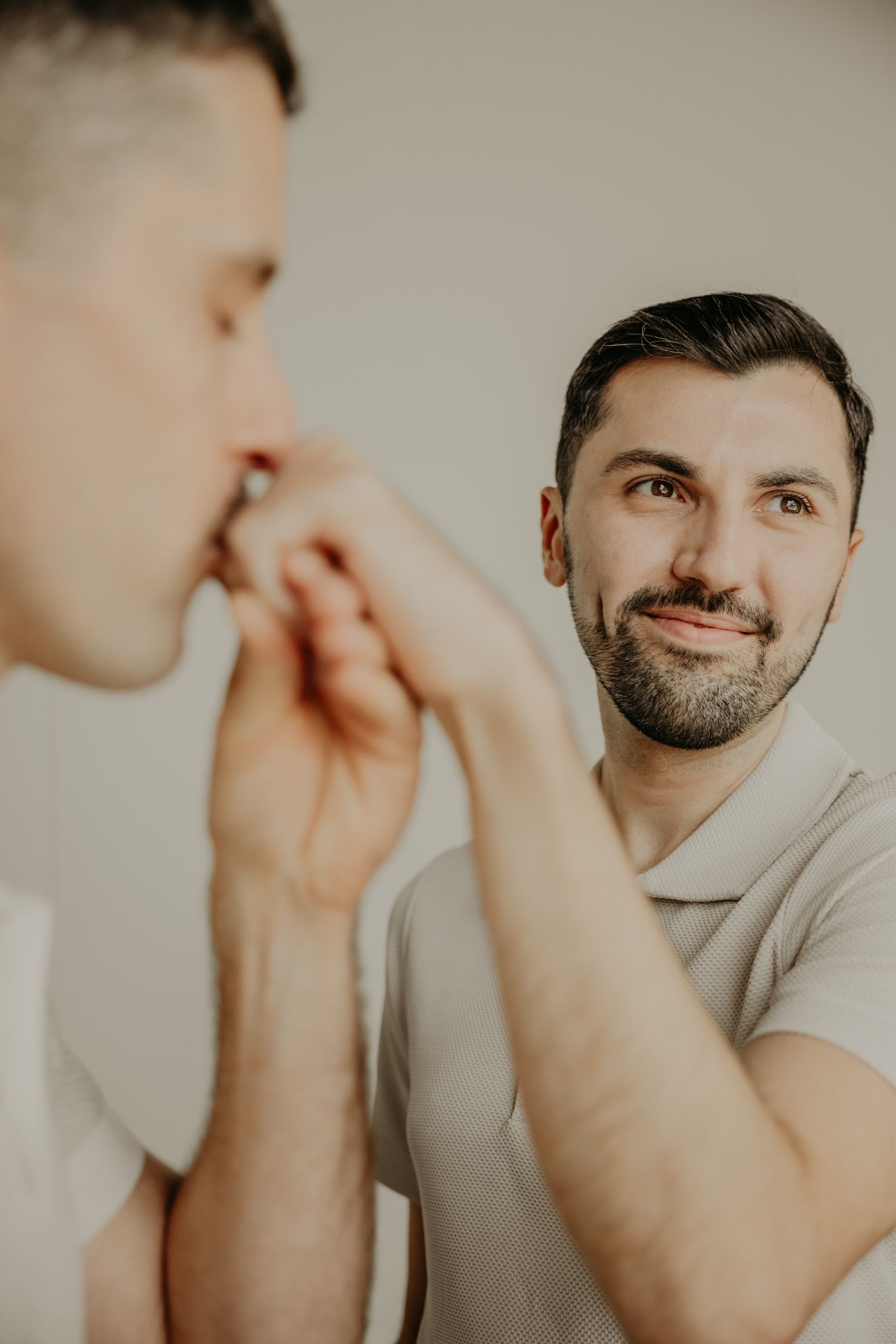 A bearded man smiles warmly while kissing another person's hand in a tender, romantic gesture indoors.