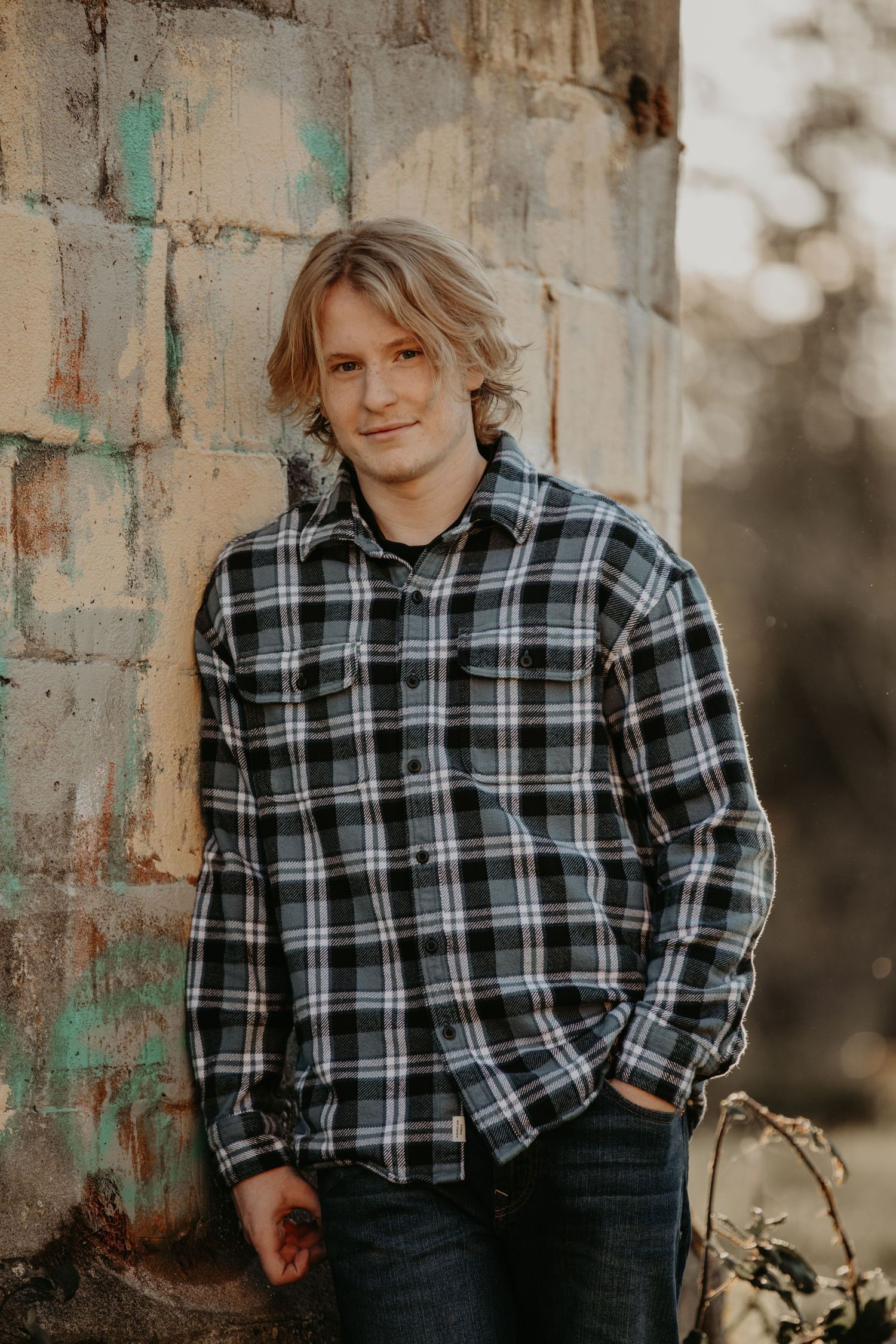 Someone in a black and white plaid flannel shirt and jeans poses casually against a concrete wall outdoors.