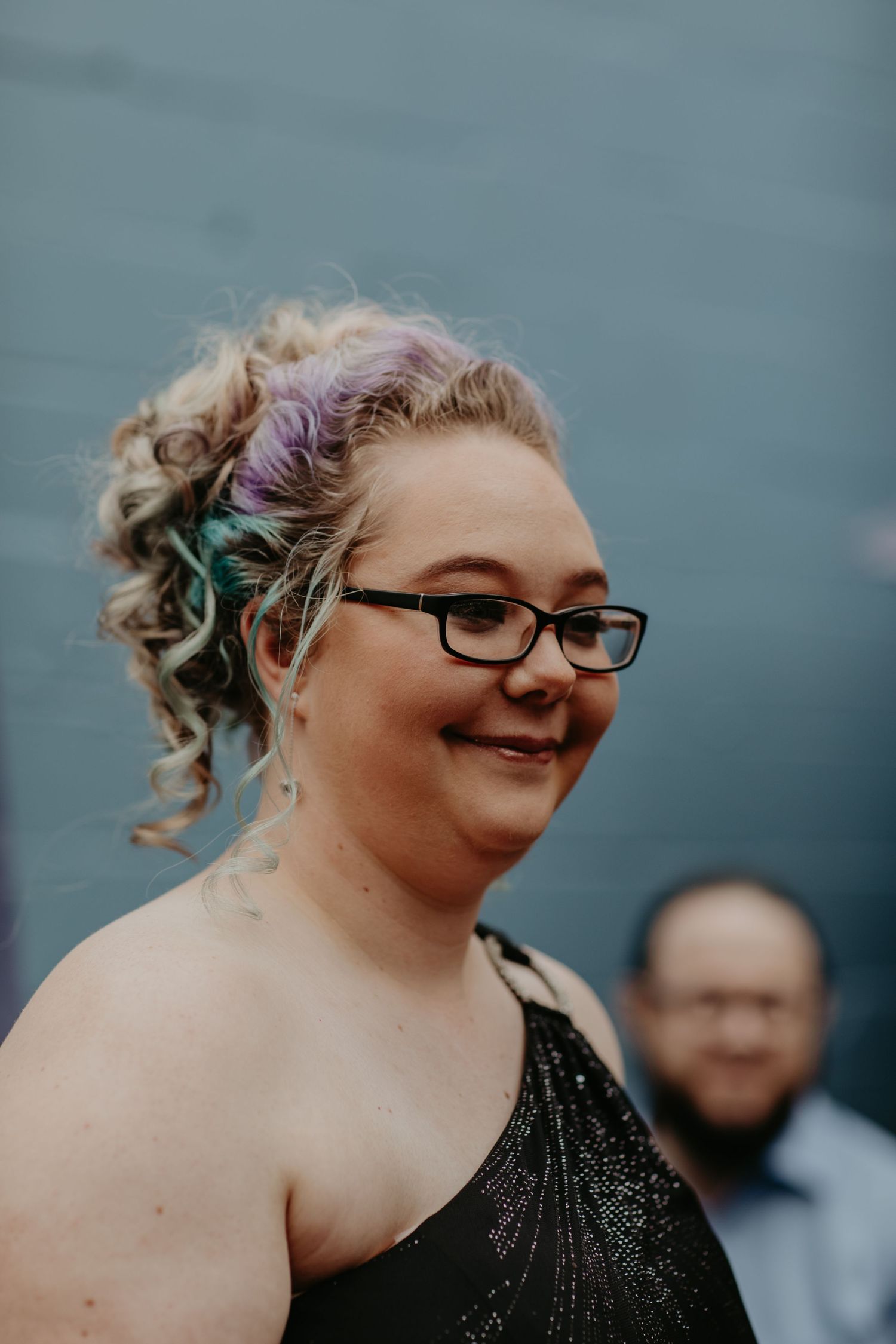 A smiling person with glasses wearing a sparkly black formal dress and curled hair with colorful streaks.