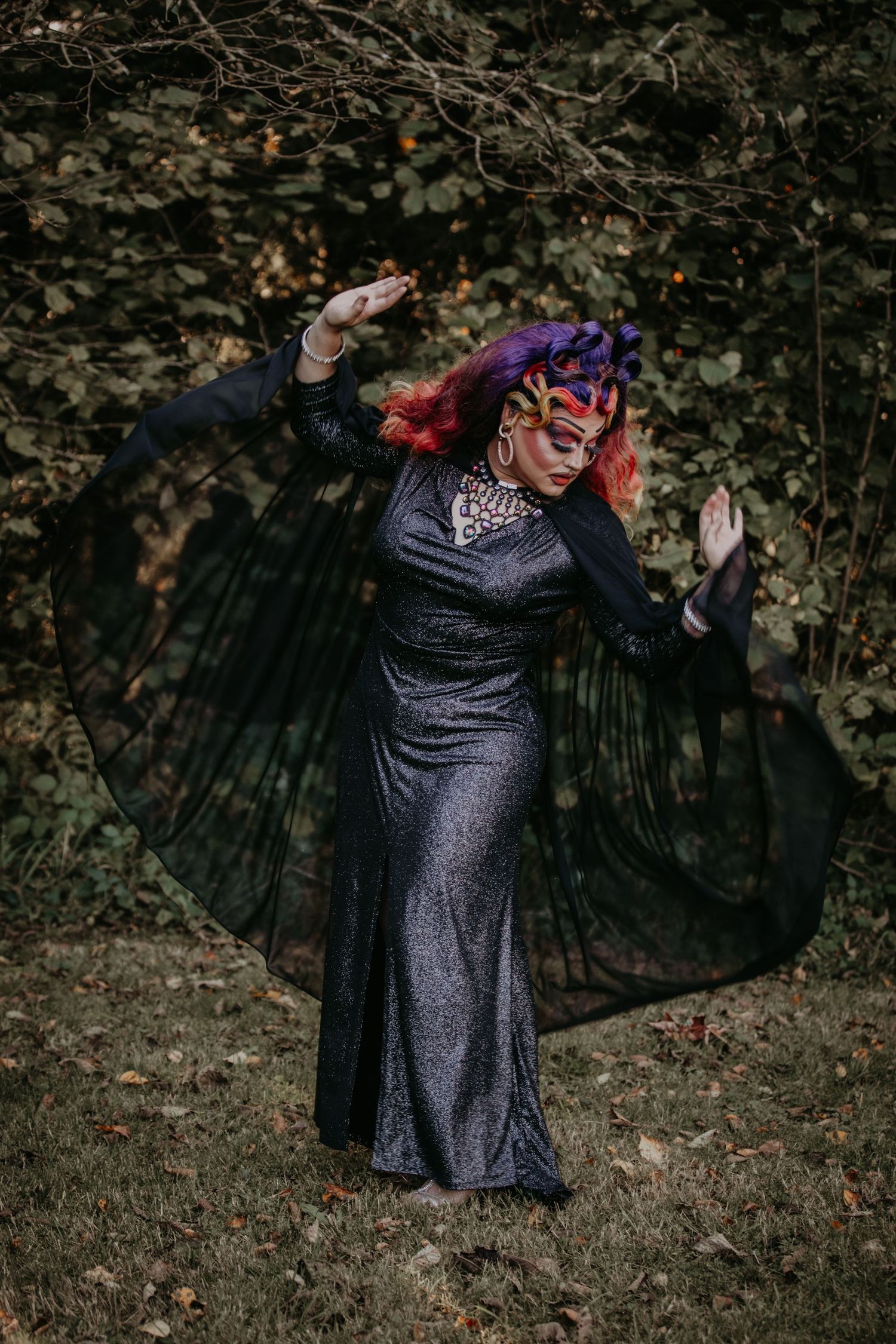 A person in a sparkling black gown and purple hair strikes a dramatic pose in an outdoor garden setting.