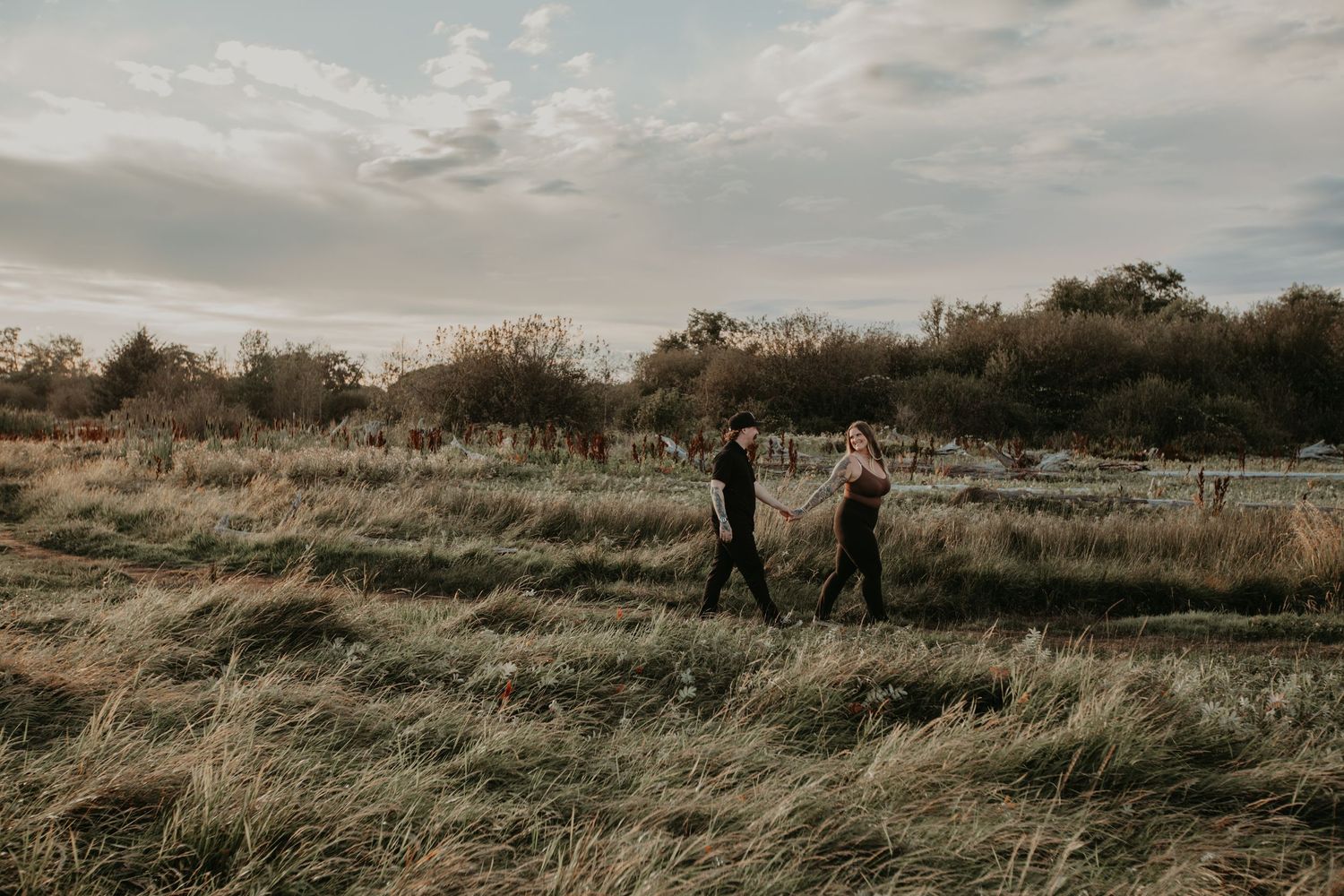 Two people walk through tall grass in a moody autumn field at golden hour with silhouetted trees in the background.