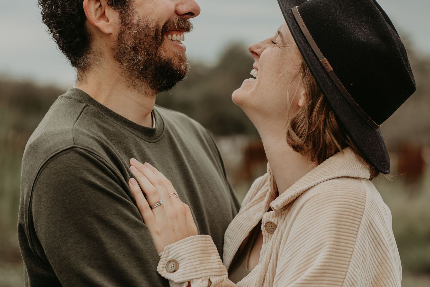 A couple shares a tender moment outdoors, with one wearing a dark hat and cream sweater, the other in olive green.