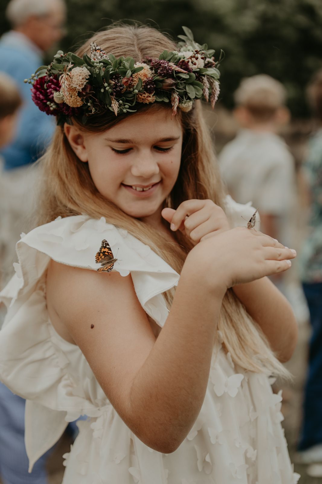 A young girl in a white dress wearing a floral crown delightfully interacts with a butterfly.