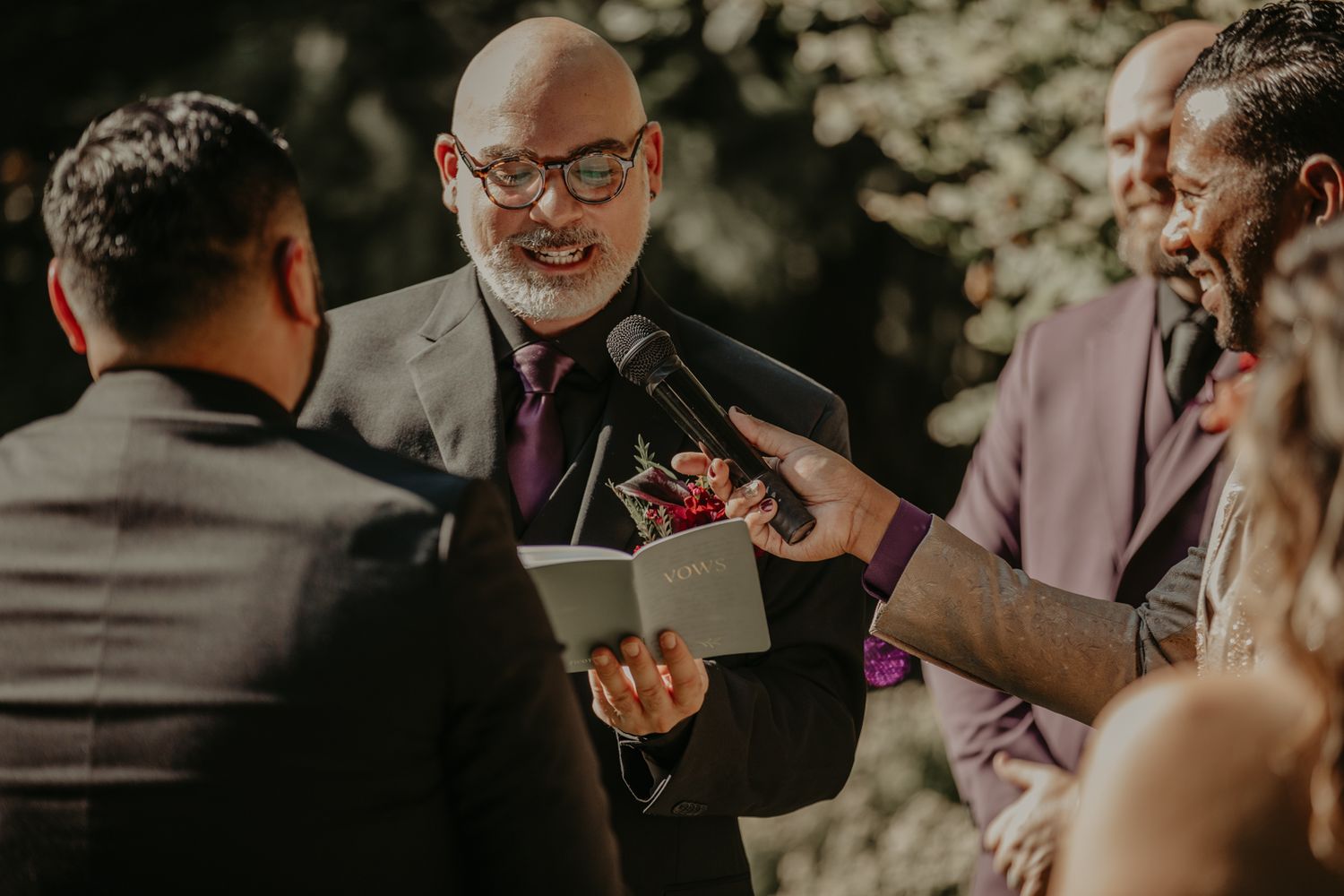 An officiant in glasses and a suit conducts an outdoor wedding ceremony while holding a tablet in natural lighting.